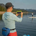 Waterproof Pak in Heather Blue with a roll top closure and zippered second compartment displayed around the waist of someone taking a picture of a stand-up paddleboarder.