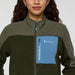 Person wearing a green and black jacket with a blue pocket featuring the Cotopaxi logo on a white background.
