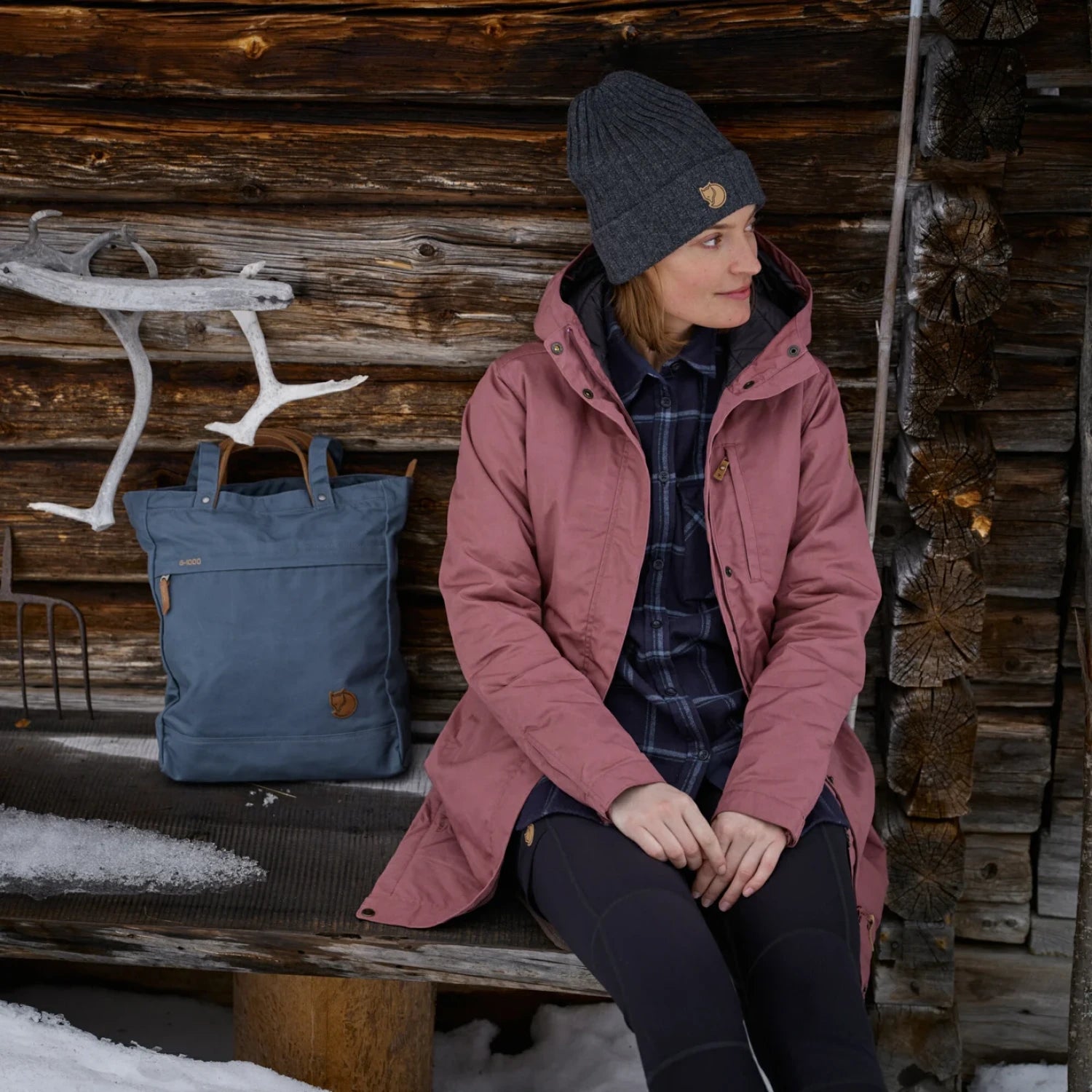 fjallraven womens kiruna padded parka jacket in port color front view worn by model sitting on a wooden bench near snow