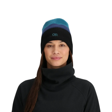 Woman wearing a black turtleneck and a striped beanie with a brand logo on a light gray background