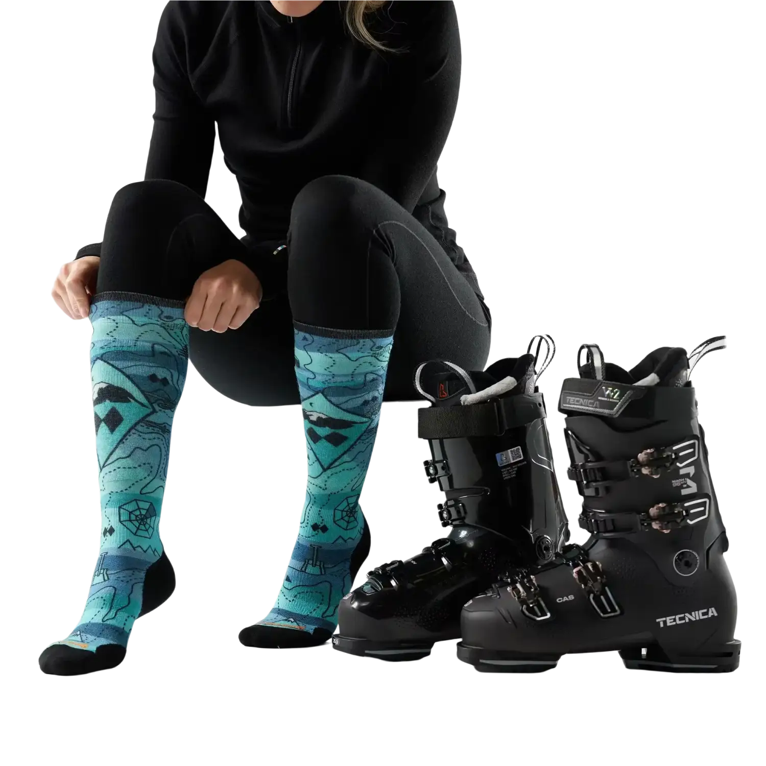 Person wearing black ski boots with blue patterned socks, standing next to two pairs of black ski boots on a white background.