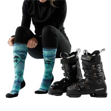 Person wearing black ski boots with blue patterned socks, standing next to two pairs of black ski boots on a white background.
